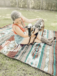 Woman in denim shorts and cowgirl boots sitting on a Southwestern blanket in a sunny backyard, gently petting a brown-and-white goat.