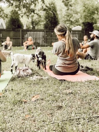 Outdoor goat yoga class on a grassy backyard lawn — woman with a ponytail seated on a pink mat while two small goats nuzzle near her feet and other participants relax on mats in the background.