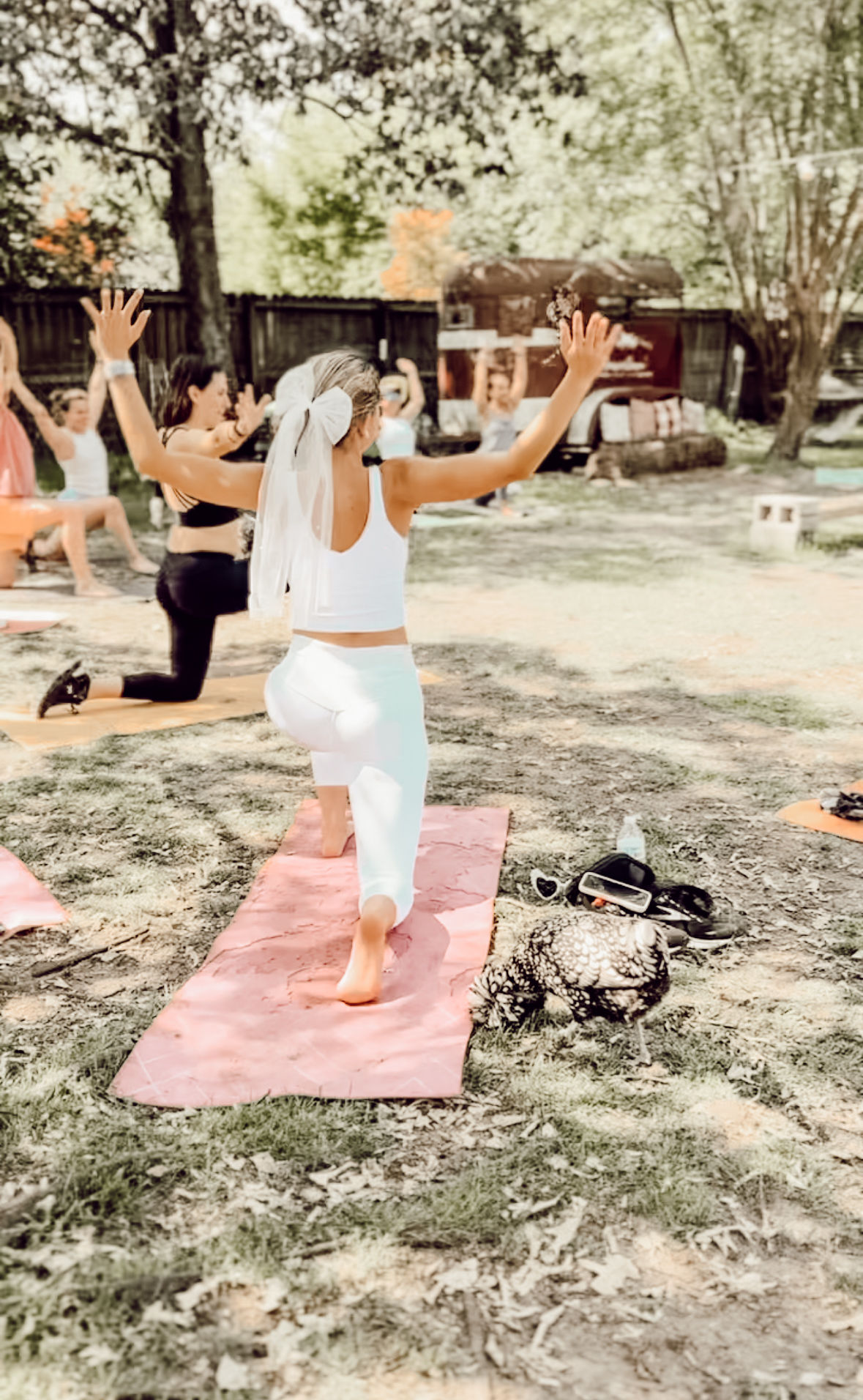 Outdoor yoga class on grass with women on mats; a participant in white leggings and a bow-like veil lunges on a pink mat with arms raised, trees and a wooden fence in the background.