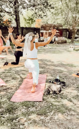 Outdoor yoga class on grass with women on mats; a participant in white leggings and a bow-like veil lunges on a pink mat with arms raised, trees and a wooden fence in the background.