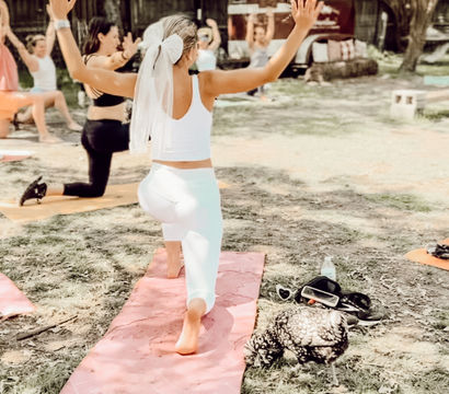 Outdoor yoga class on grass with women on mats; a participant in white leggings and a bow-like veil lunges on a pink mat with arms raised, trees and a wooden fence in the background.