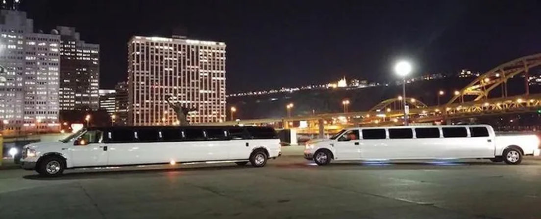 Two white stretch limousines parked on a waterfront at night with an illuminated city skyline, arched bridge and bright moon in the background.