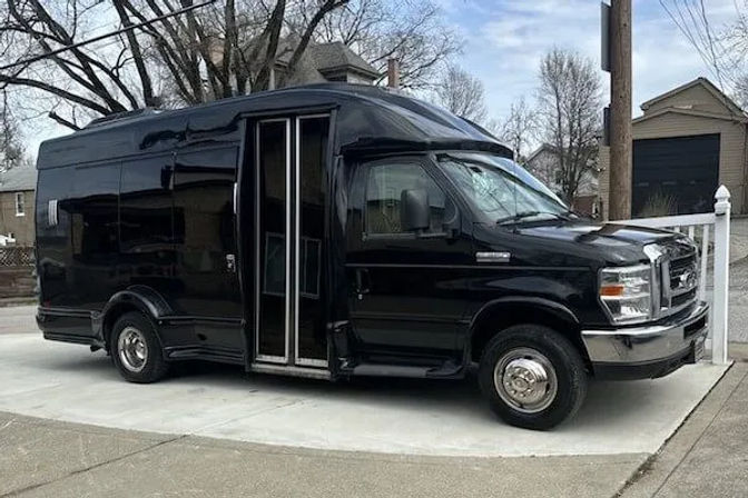 Sleek black passenger shuttle van with folding double doors parked on a concrete driveway in a suburban neighborhood with houses and bare trees in the background