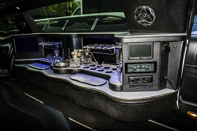 Sleek stretch limousine interior featuring a curved built-in bar with crystal decanters, martini glasses, LED ambient lighting, cup holders and plush leather seating—perfect for nightlife rides.