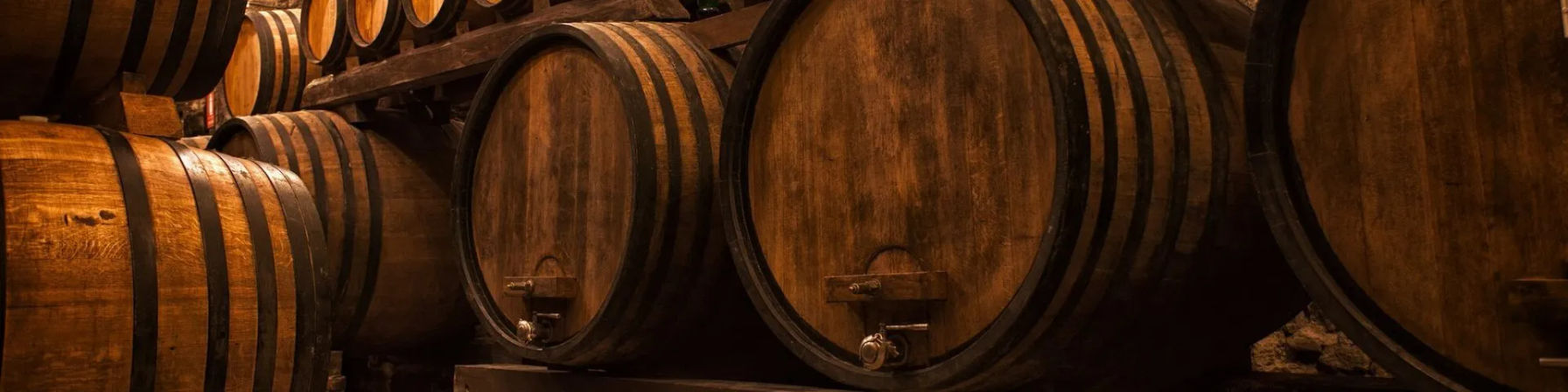 Dimly lit wine cellar with large oak barrels stacked on wooden racks, warm amber lighting highlighting aging casks.