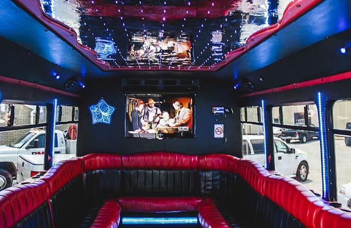 Party bus interior with red-and-black wraparound leather seating, blue LED accent lights, starry mirrored ceiling and a wall-mounted TV, parked vehicles visible through windows