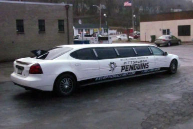 White stretch limousine with Pittsburgh Penguins hockey graphics parked in a small-town parking lot beside brick buildings and an American flag