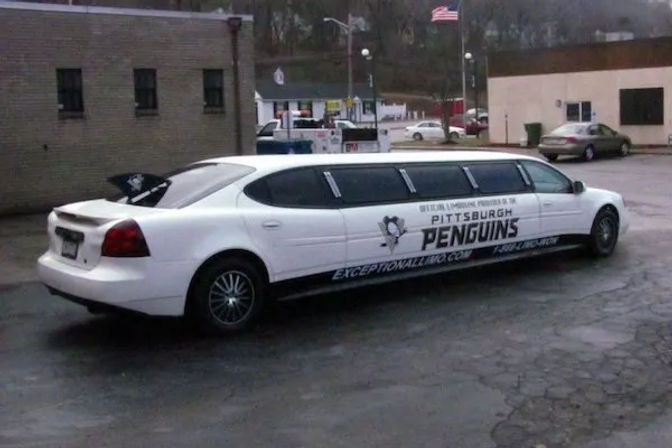 White stretch limousine with Pittsburgh Penguins hockey graphics parked in a small-town parking lot beside brick buildings and an American flag