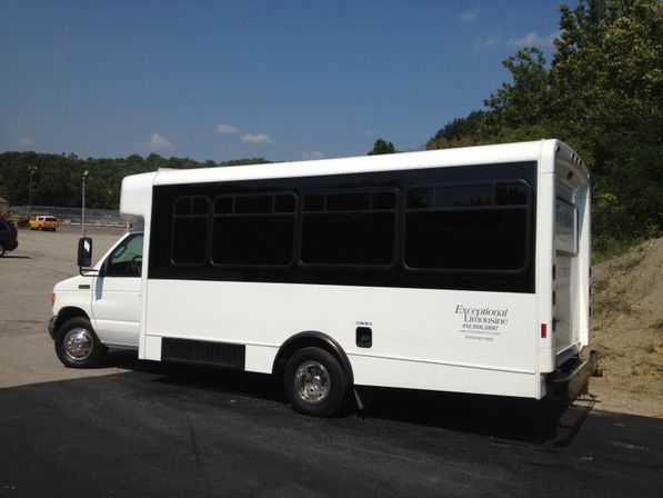 White passenger shuttle bus with dark-tinted windows parked in a suburban asphalt lot beside a wooded hillside under a clear blue sky