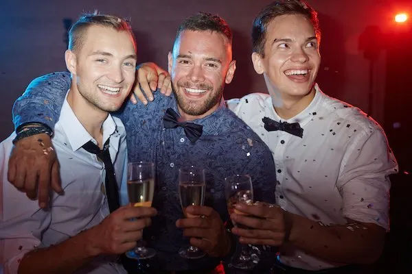 Three smiling men in dress shirts and bow ties toasting champagne amid confetti at a lively downtown nightclub party.