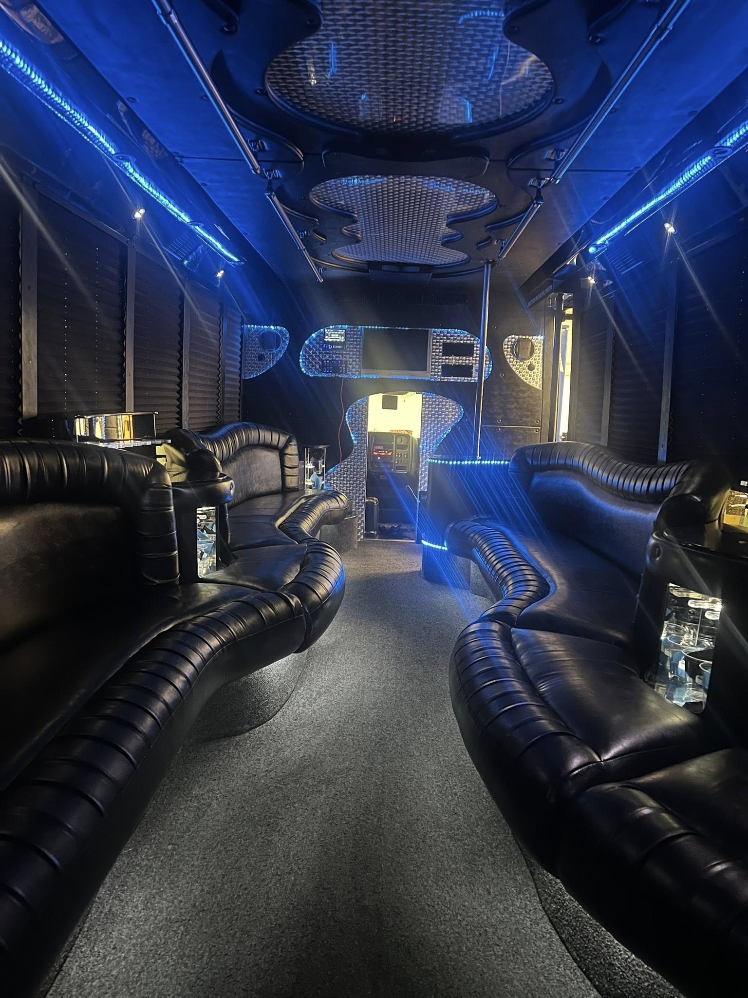 Sleek luxury party bus interior with curved black leather bench seating, blue LED accent lighting, mirrored panels, a dance pole, and a carpeted center aisle.