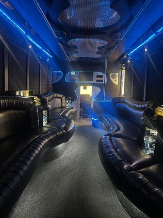 Sleek luxury party bus interior with curved black leather bench seating, blue LED accent lighting, mirrored panels, a dance pole, and a carpeted center aisle.