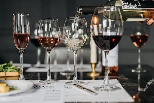 Wine tasting flight on a marble bar: four stemmed glasses with rosé, white being poured and red wine, tasting notes and a small cheese plate at a wine bar.