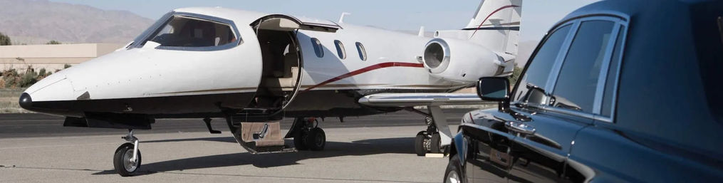 White private jet with open door parked on tarmac beside a glossy black luxury sedan at a small desert airport with mountains in the background