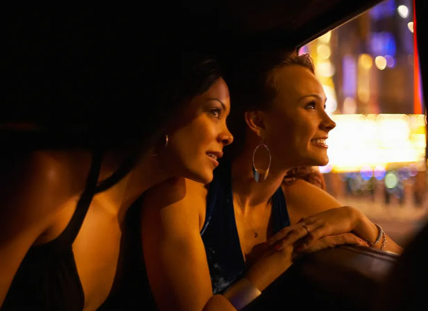 Two friends in evening dresses leaning out of a car window at night, smiling and taking in colorful downtown nightlife lights