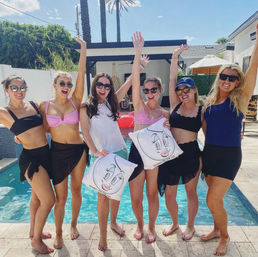 Six friends in swimsuits and sunglasses striking poses with arms raised at a sunny backyard pool party with palm trees, two holding cartoon-face pillows.