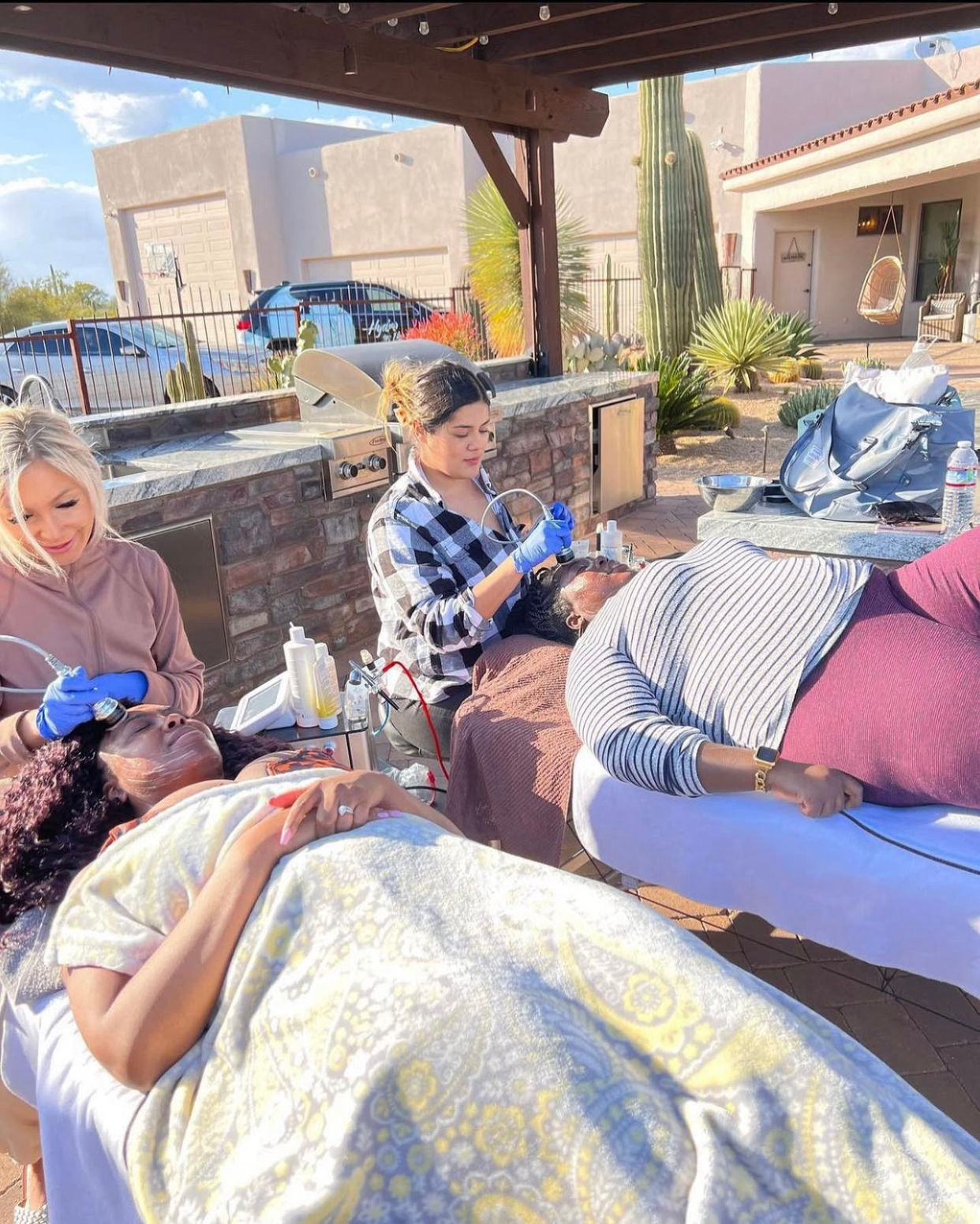 Two clients reclining on spa beds while estheticians perform outdoor facial treatments under a wooden pergola in a sunny desert backyard with saguaro cactus and a southwestern-style house nearby.