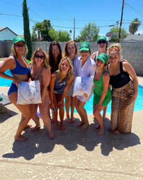Nine friends smiling and posing at a sunny backyard pool party, wearing swimwear and green caps and holding cartoon-illustrated pillows, with palm trees, a concrete pool deck and cinderblock wall in the background.