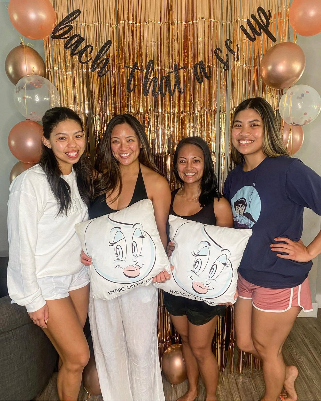 Four smiling friends posing in a living-room bachelorette celebration in front of a rose-gold fringe backdrop and balloons, holding cartoon-face throw pillows.