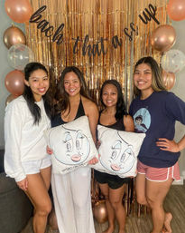Four smiling friends posing in a living-room bachelorette celebration in front of a rose-gold fringe backdrop and balloons, holding cartoon-face throw pillows.
