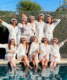 Ten women in white robes and pink heart-shaped sunglasses posing poolside with their feet in the water, playful bachelorette/bridal party at a sunny backyard pool