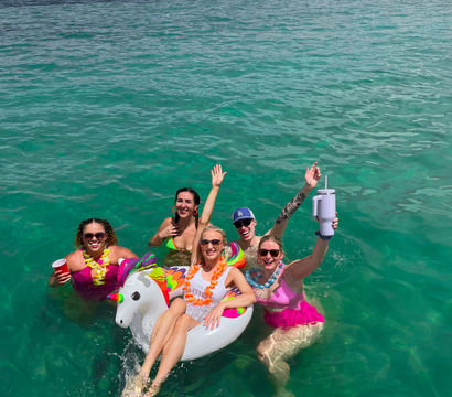 Group of friends partying in clear turquoise water near a mangrove-lined shore, one lounging on a colorful unicorn float while others hold drinks and wear leis — tropical boat celebration.