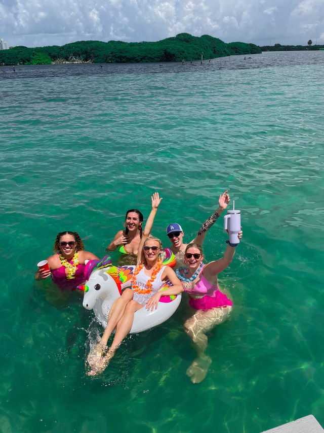 Group of friends partying in clear turquoise water near a mangrove-lined shore, one lounging on a colorful unicorn float while others hold drinks and wear leis — tropical boat celebration.