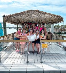 Group of friends at a waterfront marina tiki hut on a dock — women in heart-shaped sunglasses and colorful leis lift a bride in a white dress for a festive bachelorette photo.