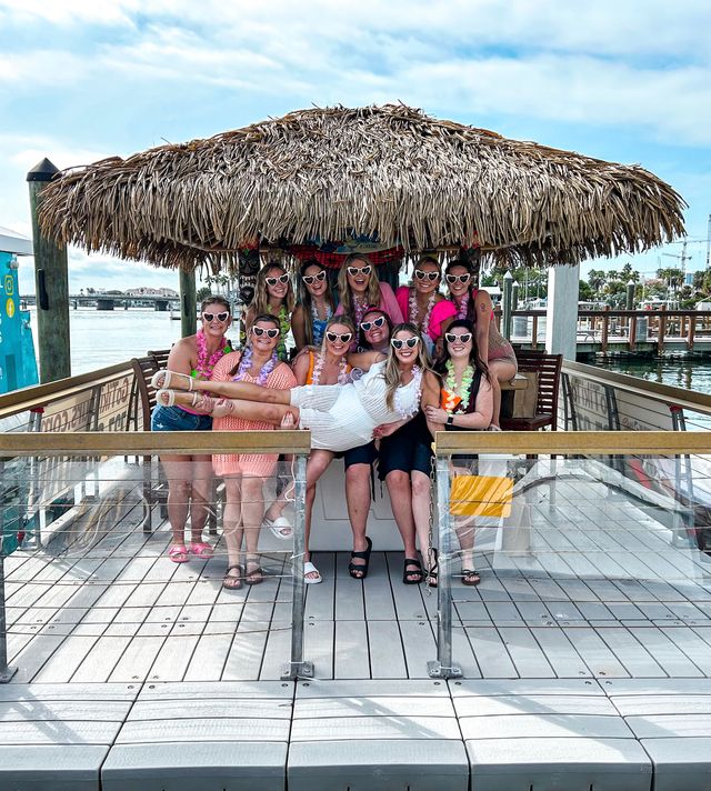 Group of friends at a waterfront marina tiki hut on a dock — women in heart-shaped sunglasses and colorful leis lift a bride in a white dress for a festive bachelorette photo.