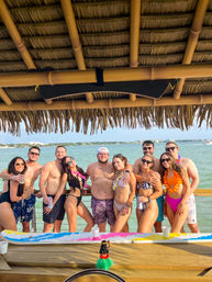 Smiling group of friends in swimwear and leis posing under a thatched tiki hut on a sunny coastal pier with the ocean and distant shoreline behind them