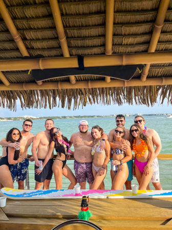 Smiling group of friends in swimwear and leis posing under a thatched tiki hut on a sunny coastal pier with the ocean and distant shoreline behind them