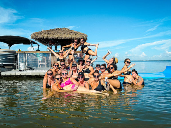 Group of women in swimsuits posing and laughing in shallow water beside a tiki-roofed dock and boat under a bright blue summer sky