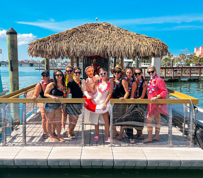 Group of women in sunglasses and colorful leis posing under a thatched tiki palapa on a sunny marina dock with boats, blue sky and an inflatable groom prop