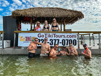 Waterfront tiki hut on a dock with a group of friends — three women under a thatched palapa and several men waist‑deep in the water holding drinks under a cloudy blue sky.