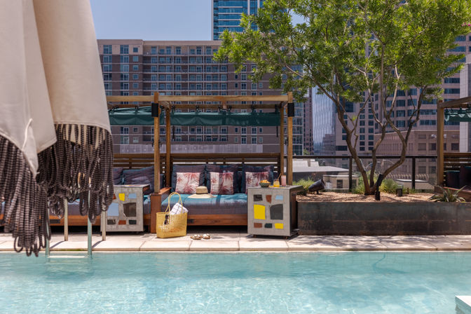 Sunny rooftop pool with turquoise water, shaded cabana lounge featuring patterned cushions, a straw tote and sandals on the deck, fringe umbrella at the edge and urban skyscrapers in the background
