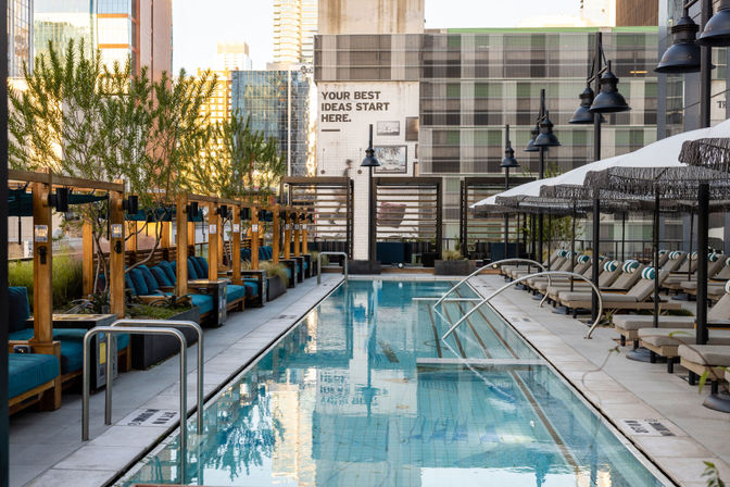 Rooftop urban oasis with a long blue-tiled lap pool, wooden cabanas with teal cushions on the left and cushioned loungers with fringe umbrellas on the right, city high-rises reflecting in the water.
