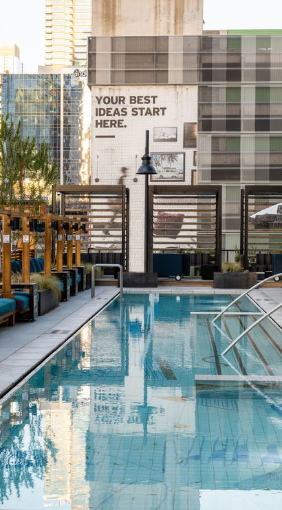 Rooftop urban oasis with a long blue-tiled lap pool, wooden cabanas with teal cushions on the left and cushioned loungers with fringe umbrellas on the right, city high-rises reflecting in the water.