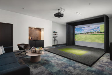 Contemporary game room with indoor golf simulator — ceiling projector and large course screen, turf hitting mat, golf clubs, lounge seating and snack table.