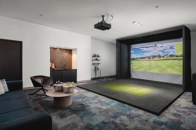 Contemporary game room with indoor golf simulator — ceiling projector and large course screen, turf hitting mat, golf clubs, lounge seating and snack table.
