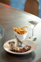 Bistro-style plate of sliced seared steak medallions topped with herb butter, seasoned French fries in a silver cup with dipping sauce, and a lemon-twist martini on a restaurant table.