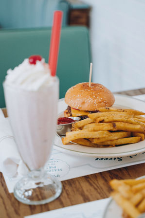 Classic diner meal: cheeseburger with sesame bun and seasoned fries on a plate, paired with a strawberry milkshake topped with whipped cream and a cherry.
