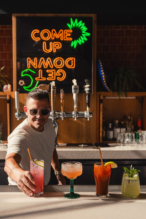 Sunlit bar bartender hands a pink cocktail across a counter with four colorful mixed drinks, draft taps and a neon "COME UP GET DOWN" sign