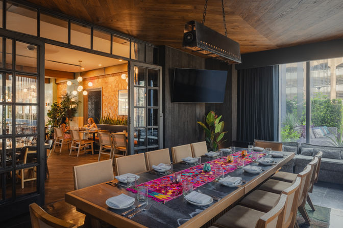 Modern private dining room in an urban restaurant with a long wooden table set for a group, vibrant embroidered table runner, beige chairs, globe pendant lights, glass partition to a cozy dining area and large windows overlooking a landscaped patio.