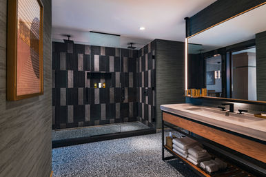 Modern luxury bathroom with glass walk-in shower of dark geometric tiles, dual rainfall heads, terrazzo floor and wood-and-stone vanity with illuminated mirror and towels.