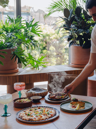 Indoor dining table spread with pizza, grilled fish, a steaming skillet of grilled meat and vegetables, bowls of salad and tortillas, and two margarita-style cocktails, lush potted plants in the background, server’s arm placing the hot skillet.