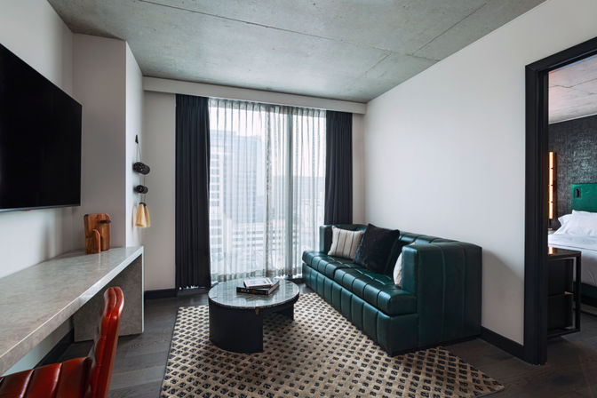 Modern hotel suite living area with emerald leather sofa, patterned rug, round coffee table and floor-to-ceiling window revealing an urban high-rise view.