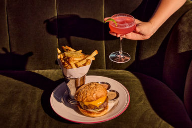 Juicy cheeseburger with melted cheddar and sesame bun, crispy fries in a metal cup with dipping sauces, and a hand holding a pink cocktail coupe with lime, served on a white plate on a green velvet restaurant booth.