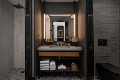 Spa-like modern hotel bathroom with illuminated rectangular mirror above stone sink and wood vanity, neatly stacked white towels on open shelf, rain shower to the left, sleek black fixtures and warm ambient lighting.