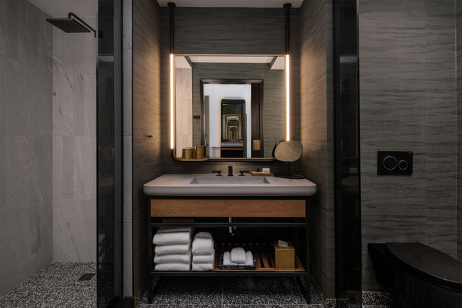 Spa-like modern hotel bathroom with illuminated rectangular mirror above stone sink and wood vanity, neatly stacked white towels on open shelf, rain shower to the left, sleek black fixtures and warm ambient lighting.