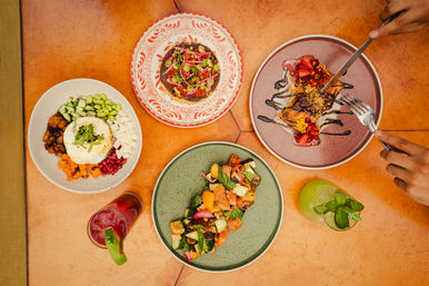 Top-down colorful brunch spread on terracotta tile table: grain bowl with sunny-side egg and diced veggies, roasted vegetable salad on a green plate, tostada with pickled onions, a dessert with strawberries and chocolate drizzle being cut, and two vibrant cocktails with lime and mint.